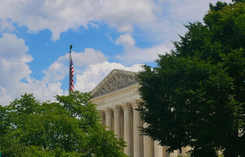 The United States Supreme Court building partially obscured by green trees, with an American flag flying on a pole against a cloudy blue sky.