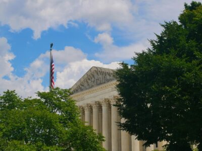 The United States Supreme Court building partially obscured by green trees, with an American flag flying on a pole against a cloudy blue sky.