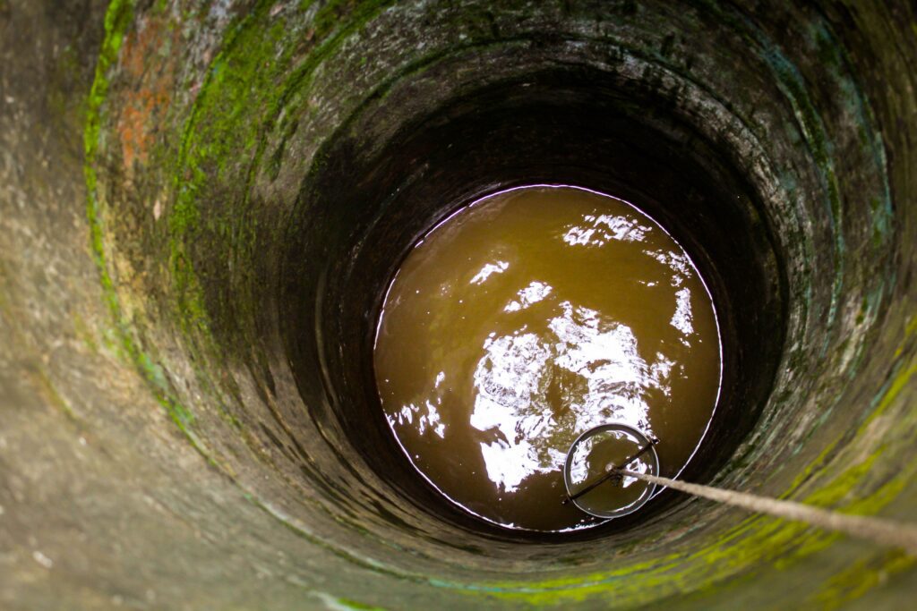 View looking down into a stone-lined well with brown water at the bottom. A bucket attached to a rope is partially submerged, and green moss is visible on the inner wall of the well.