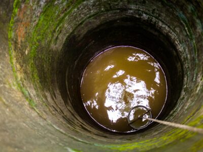 View looking down into a stone-lined well with brown water at the bottom. A bucket attached to a rope is partially submerged, and green moss is visible on the inner wall of the well.