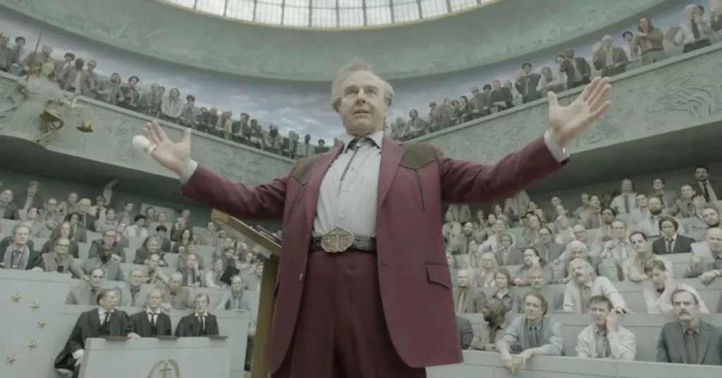 A man in a maroon suit stands with his arms outstretched before a large, seated audience in a tiered assembly hall.